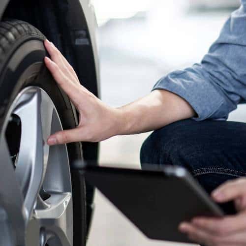 A hand holding a clipboard with another hand checking a car tyre