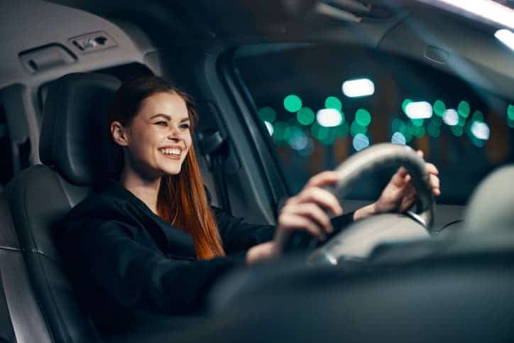 photo of a nice woman in a black shirt driving a car while sitting at the wheel with a pleasant smile on her face at night