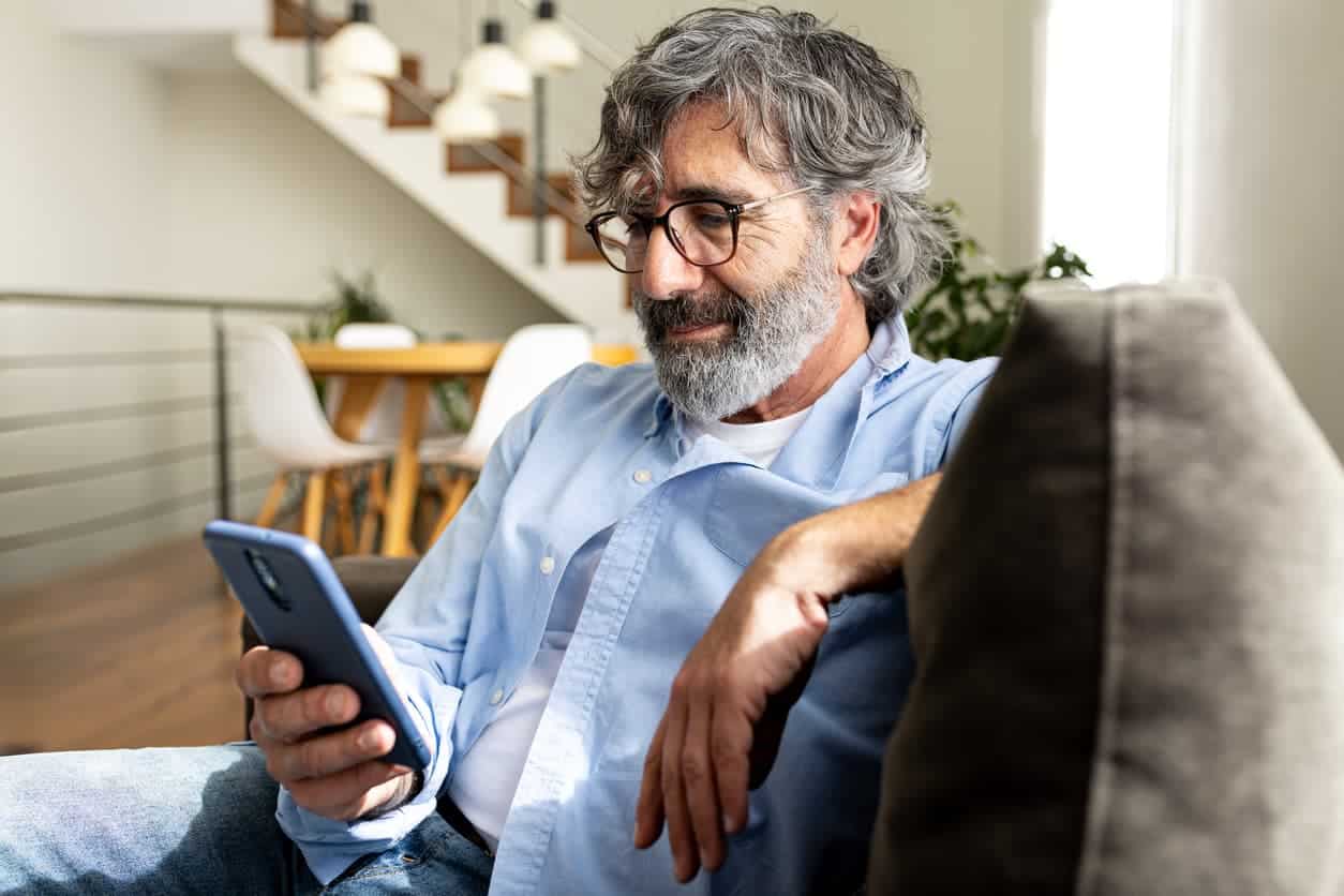 Mature man relaxing at home, sitting on the sofa sending text message with mobile phone